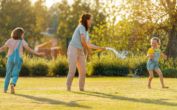 A sunny backyard in the summer as a mom is playing with her two kids with a hose.