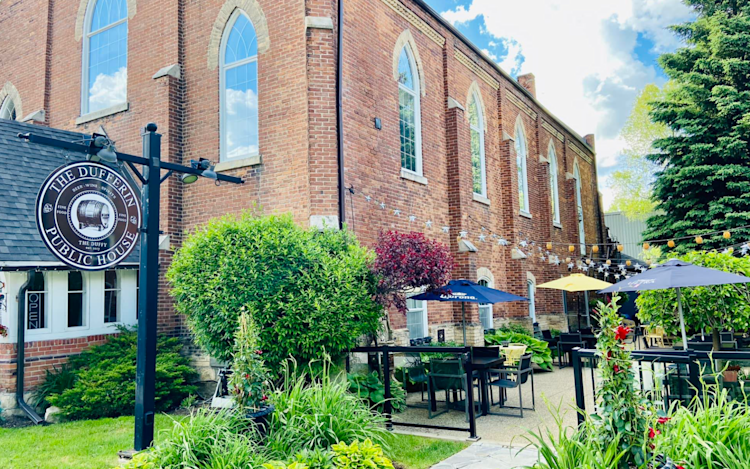 Photo of the front of the Dufferin Public House in Shelburne, Ontario. It is a converted church that was turned into a bar, and the photo shows the outdoor patio with three umbrellas and string lights.