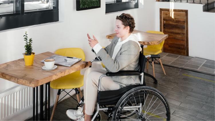 Woman in wheelchair using smartphone at a café, with her laptop and coffee in front of her.