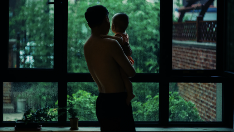 A man holding a baby looks out a window onto a forest.