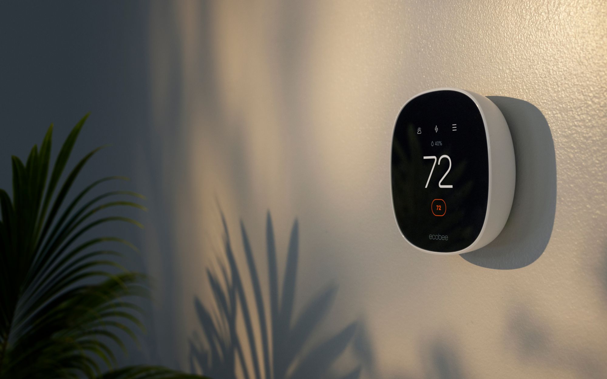 Close-up of an ecobee smart thermostat mounted on a wall, displaying 72°F in a warmly lit home with a plant nearby.