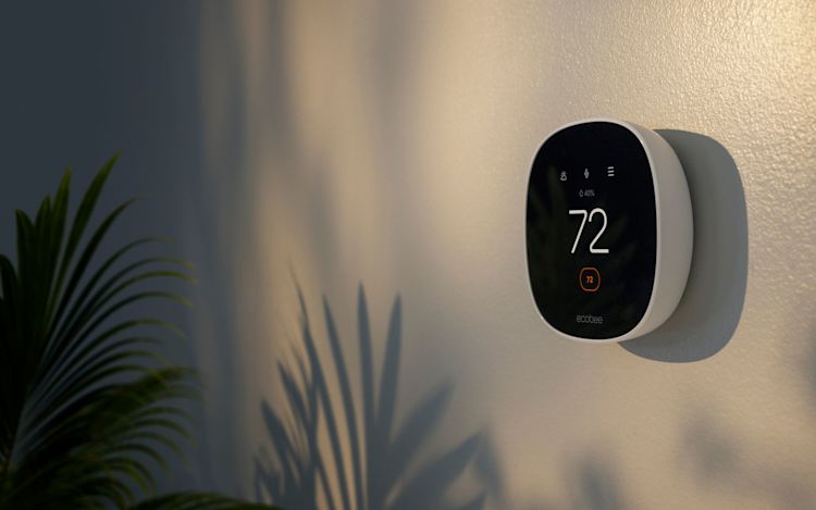 Close-up of an ecobee smart thermostat mounted on a wall, displaying 72°F in a warmly lit home with a plant nearby.