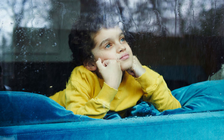 Boy on couch gazes out window on rainy day.