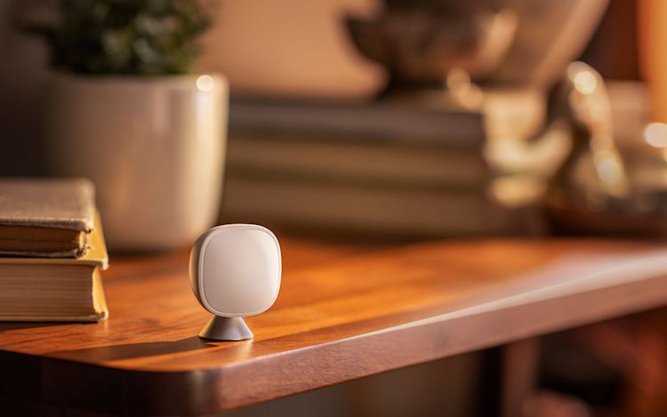 An ecobee SmartSensor sits on a wooden shelf beside a stack of books in a warmly lit living room.