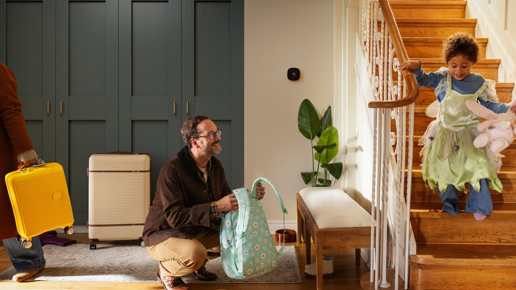 A father packs a backpack while a child comes down the stairs, with luggage by the door and an ecobee smart thermostat on the wall.