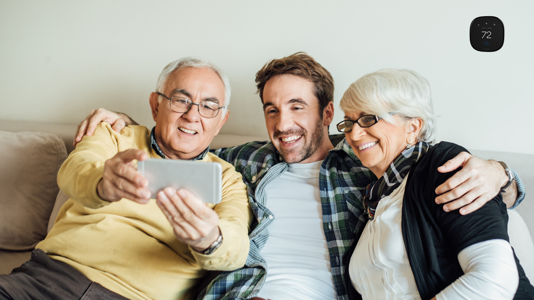 A middle-aged man with his elderly Mom and Dad on the couch and the Dad is taking a selfie of all three of them with his phone.