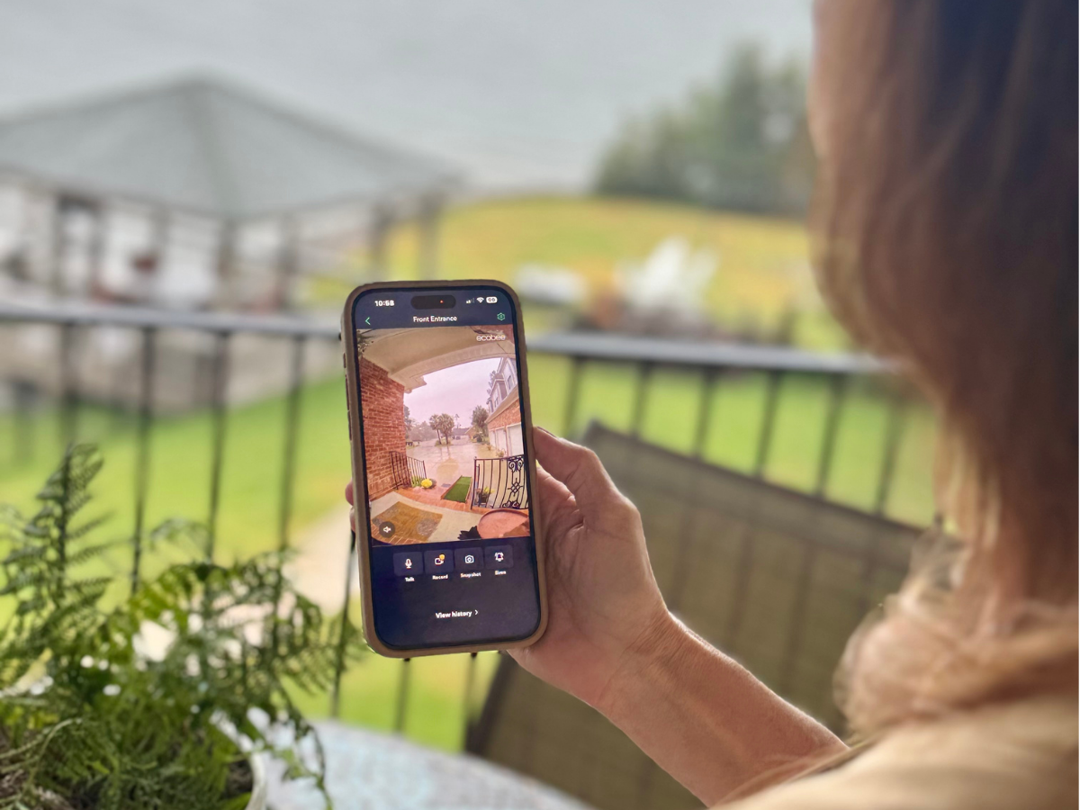 A woman is sitting on her backyard balcony holding her phone and on the screen is a front door view of ecobee's Smart Doorbell Camera with a visitor at the door. 
