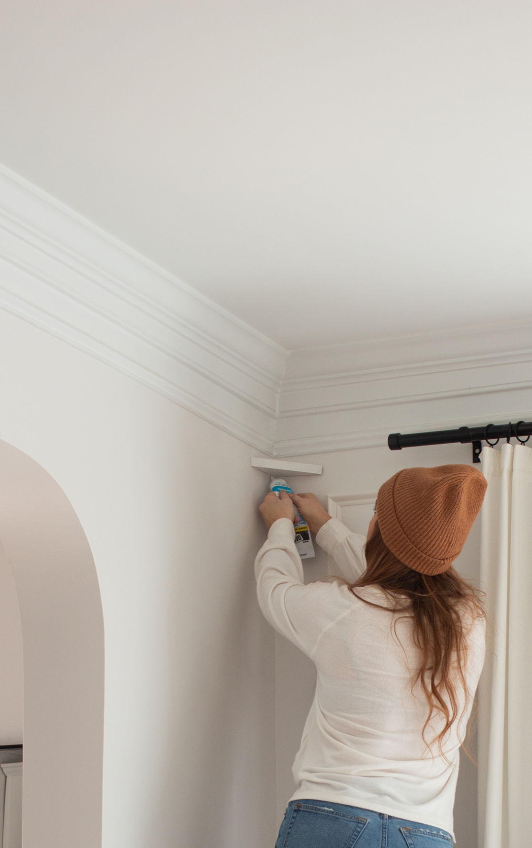 Cass from CASSMAKESHOME uses an adhesive to adhere a small white shelf to the corner of a room's ceiling.