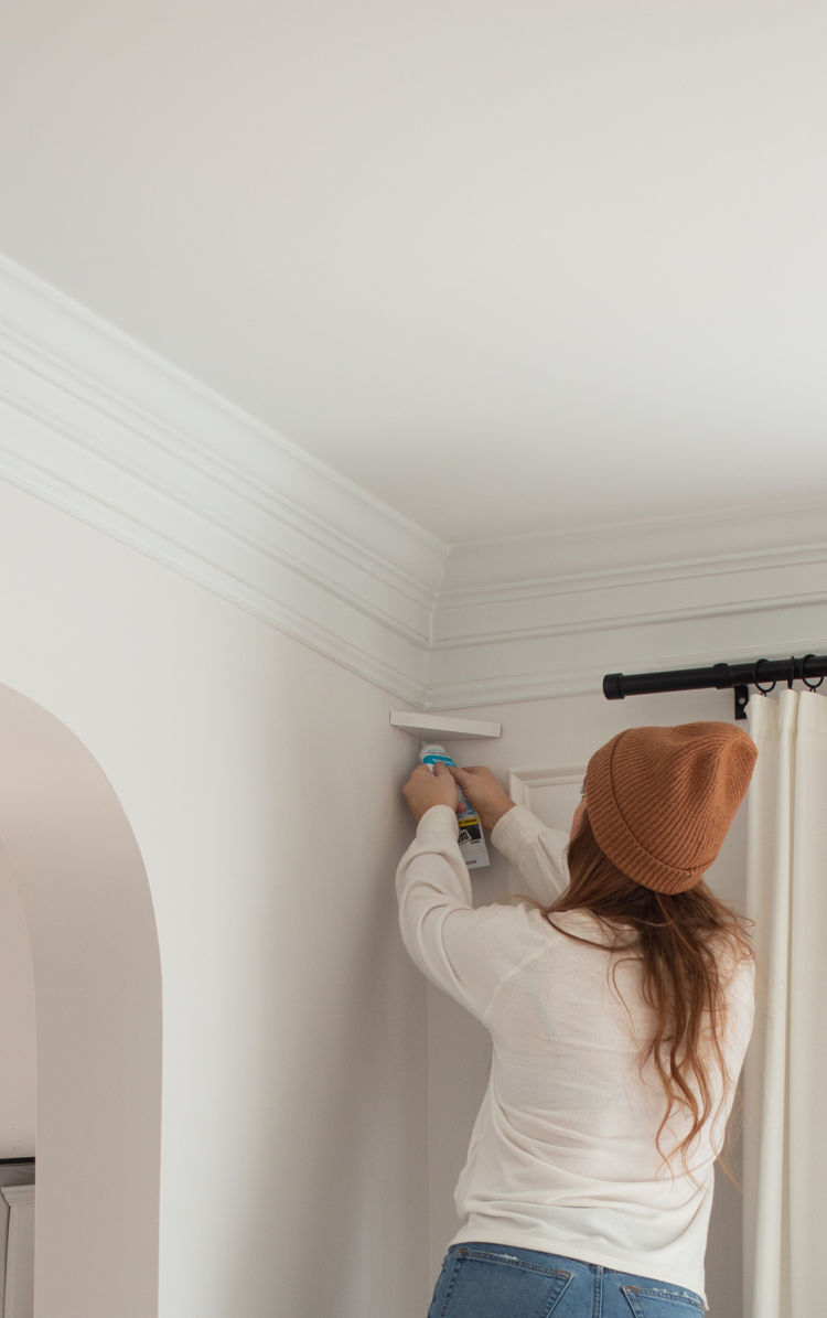 Cass from CASSMAKESHOME uses an adhesive to adhere a small white shelf to the corner of a room's ceiling.