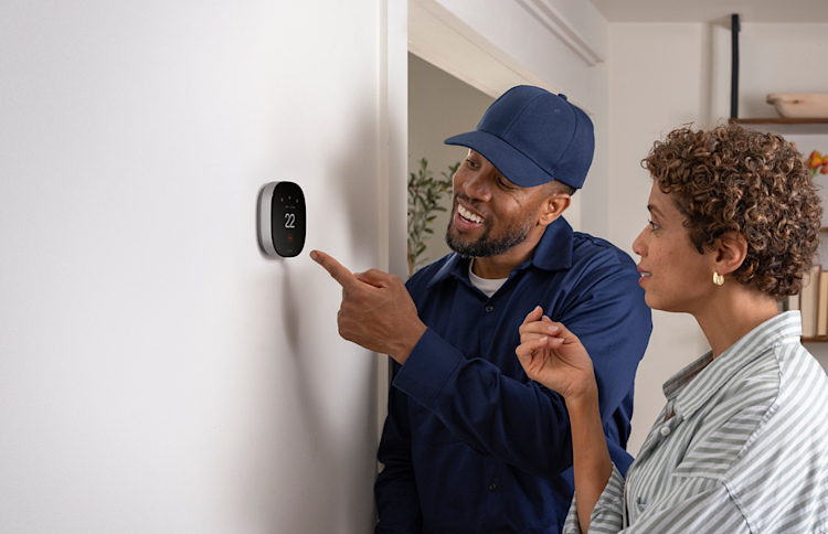 A HVAC Pro wearing a blue hat is showing a homeowner their newly installed ecobee thermostat on their white wall in their home.