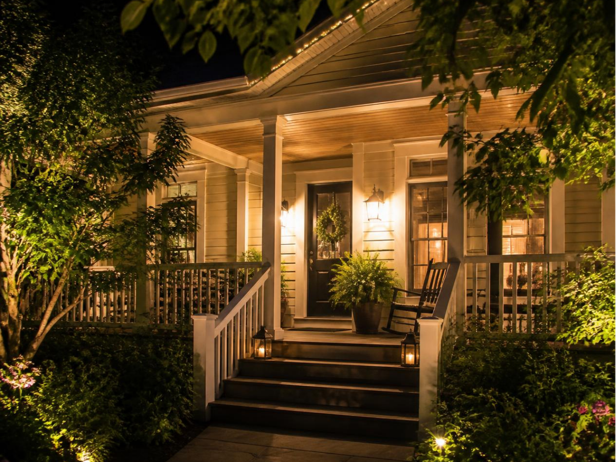 A traditional style home with greenery from trees and potted plants lit up at hight with lights and potlights with the front entrance and porch clearly visible. 