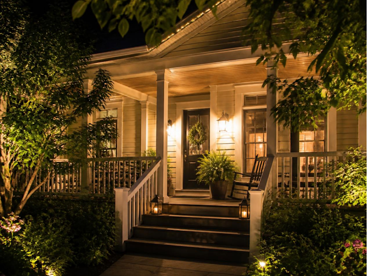 A traditional style home with greenery from trees and potted plants lit up at hight with lights and potlights with the front entrance and porch clearly visible.