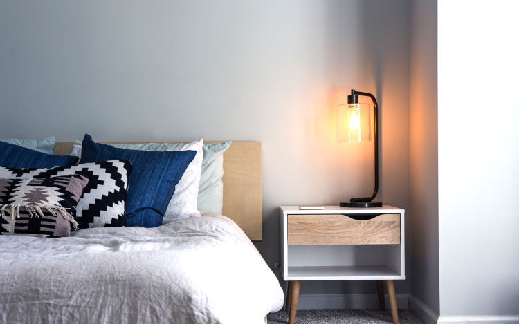 Warm temperature Edison bulb lamp sitting on modern bed side table casts a warm glow against the surrounding corner walls. 