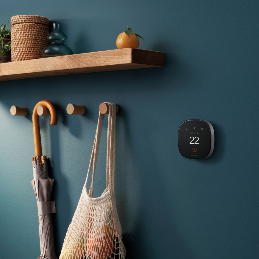 Smart Thermostat Premium attached to navy blue wall next to four wooden peg hooks, one holding an umbrella, and another holding a cotton mesh bag. A wooden shelf fashioned with home décor hangs over top, completing the scene. 