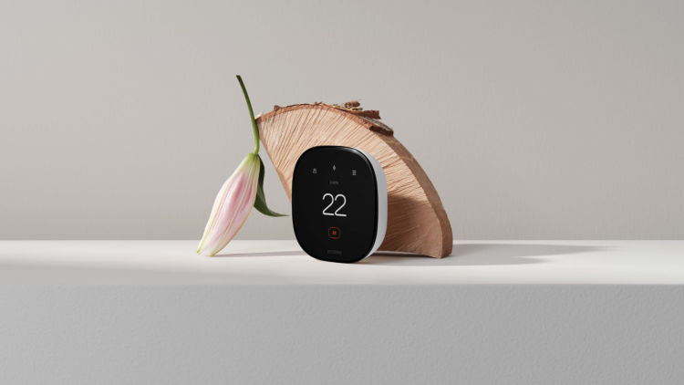 Smart Thermostat Enhanced show at a three quarter angle view sitting on a light grey surface. A piece of wood and a flower are artfully arranged behind it.