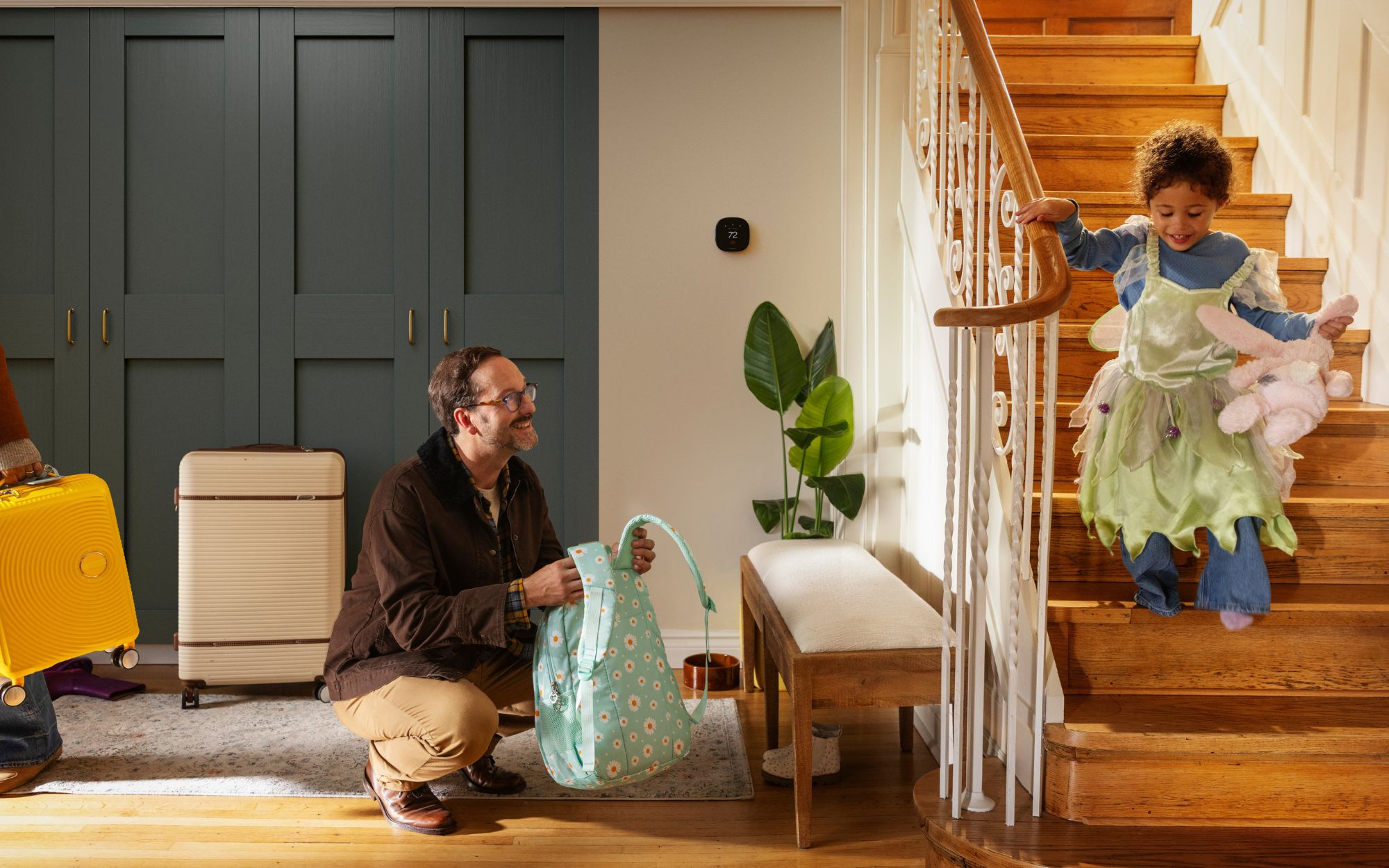 A father packs a backpack while a child comes down the stairs, with luggage by the door and an ecobee smart thermostat on the wall.