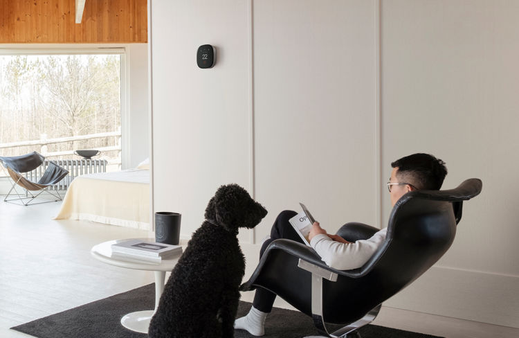 A man sits in a char reading a magazine with a black dog sitting beside him. A Smart Thermostat Premium is mounted to the wall in the background.