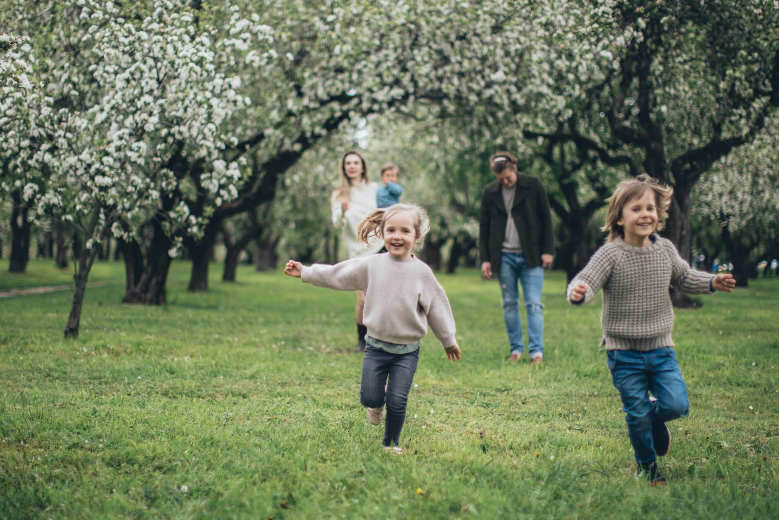 Two children running in a park with parents, baby and trees behind them.