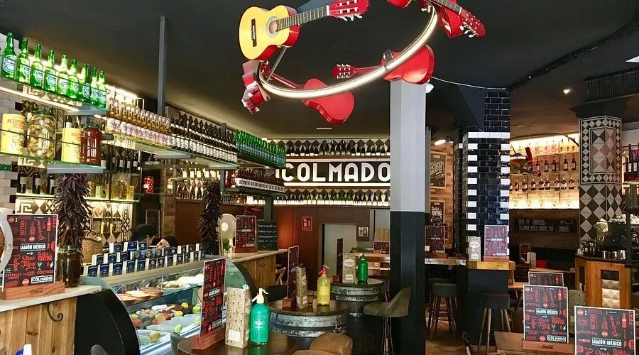 Interior de bar con decoración ecléctica, estanterías con botellas, guitarra colgante y cartel de “COLMADO” al fondo.