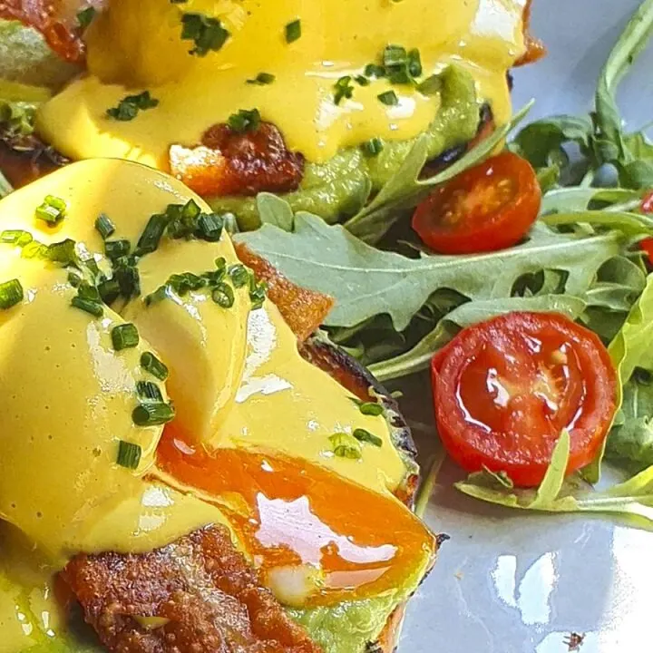 Huevos benedictinos con aguacate, bacon y salsa holandesa sobre rúcula y tomates cherry en plato blanco.