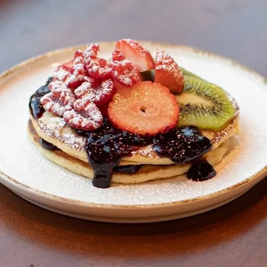 Torre de pancakes con sirope de frutos rojos, frambuesas con azúcar glas, fresas y rodajas de kiwi sobre un plato blanco con bordes dorados.