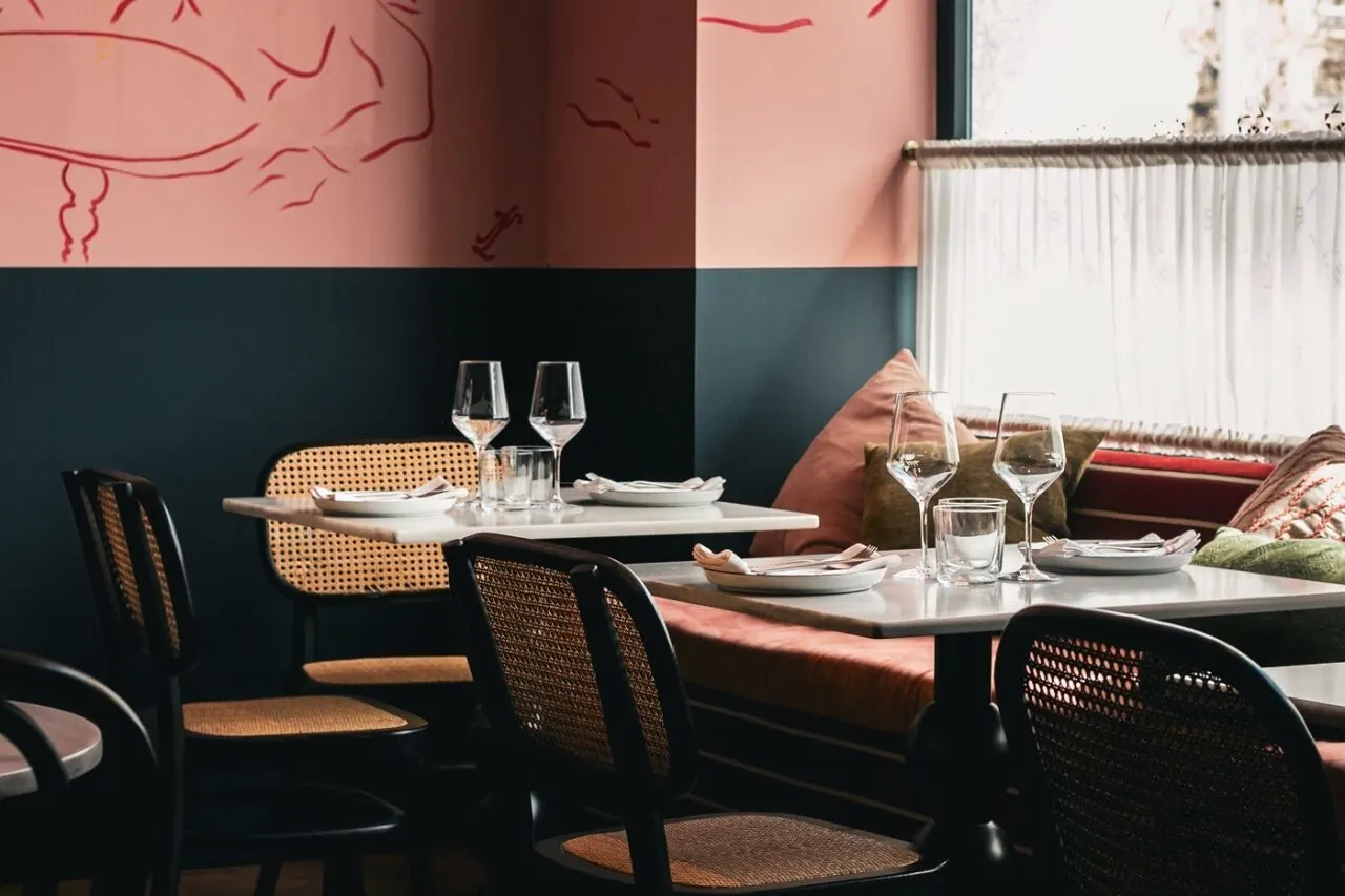 Restaurante acogedor con mesas puestas, sillas de mimbre, paredes rosa y azul con arte lineal rojo. Luz natural entra por cortinas blancas.