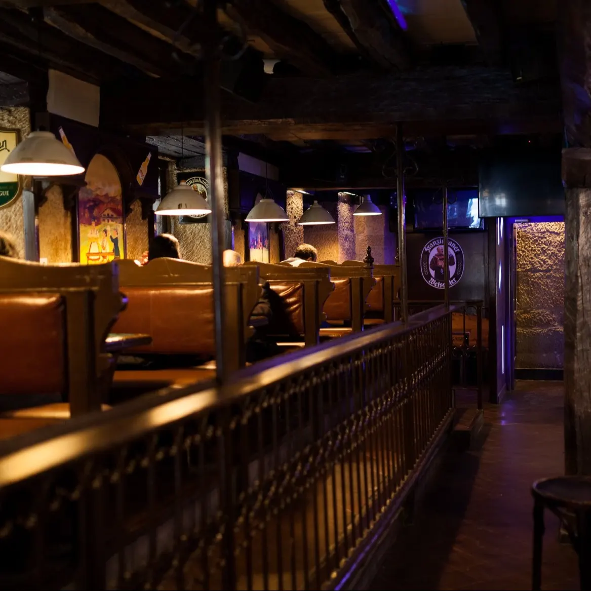 La imagen muestra el interior de Garagar, un bar con estilo rústico y acogedor. Los asientos de madera, los carteles vintage y la iluminación tenue crean un ambiente ideal para disfrutar de una cerveza en un entorno cálido y auténtico.