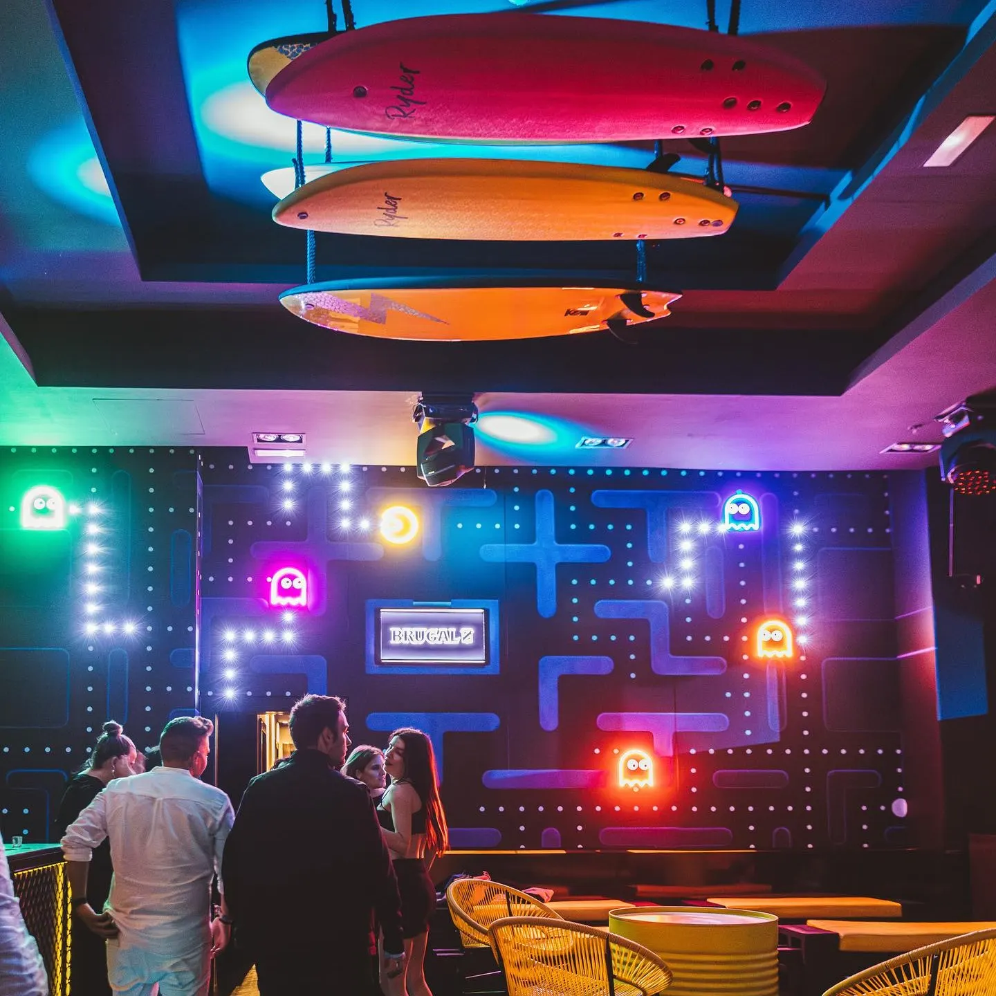 Hyp3: Bar de ambiente retro-futurista con paredes inspiradas en Pac-Man y tablas de surf en el techo. Los colores neón y la iluminación crean un espacio divertido y dinámico para los amantes del estilo arcade.