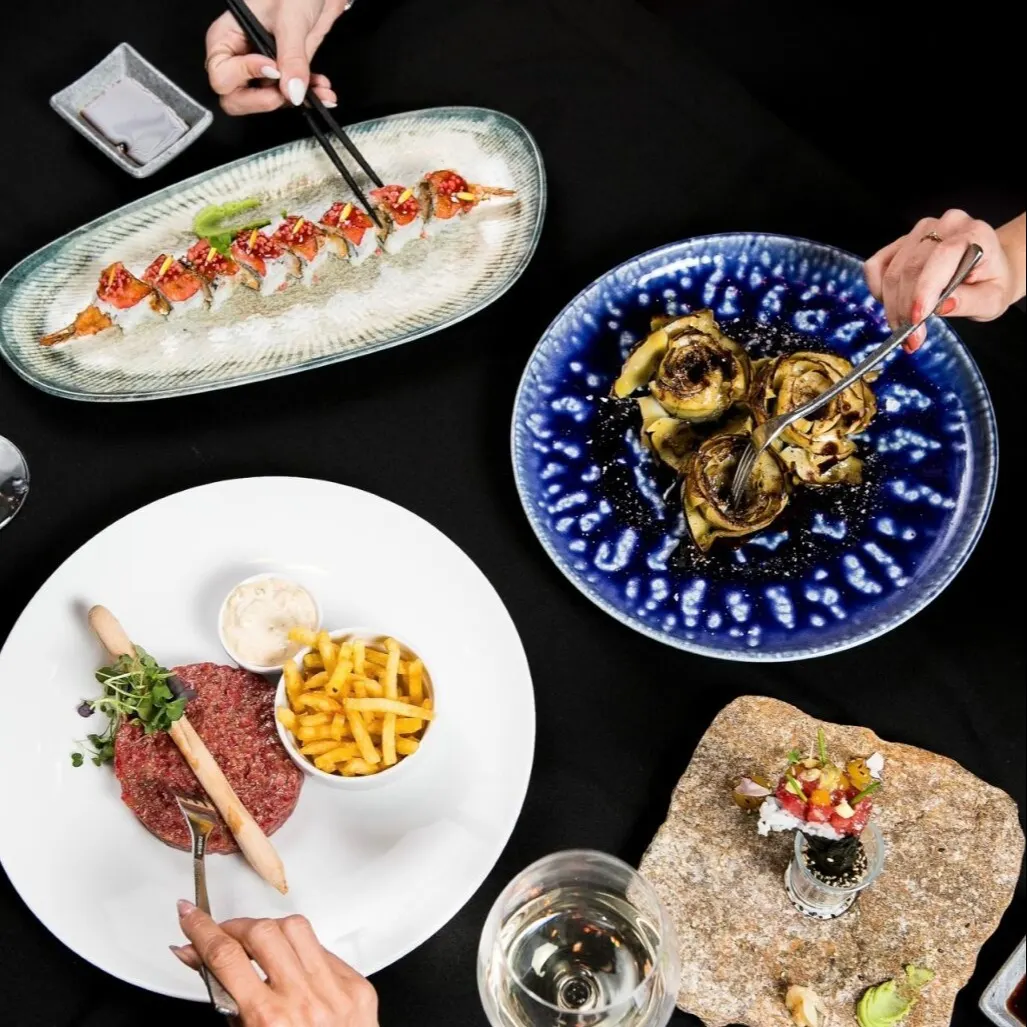 Mesa con sushi, alcachofas, steak tartar con patatas y plato sobre losa; tres manos con cubiertos y copa de vino blanco en ambiente gastronómico.