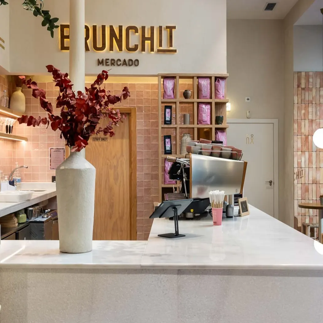 Mostrador de barra con superficie blanca, detalles en tonos tierra y rosas, y decoración floral, bajo el rótulo dorado de 'Brunchit Mercado', en un interior cálido y moderno.