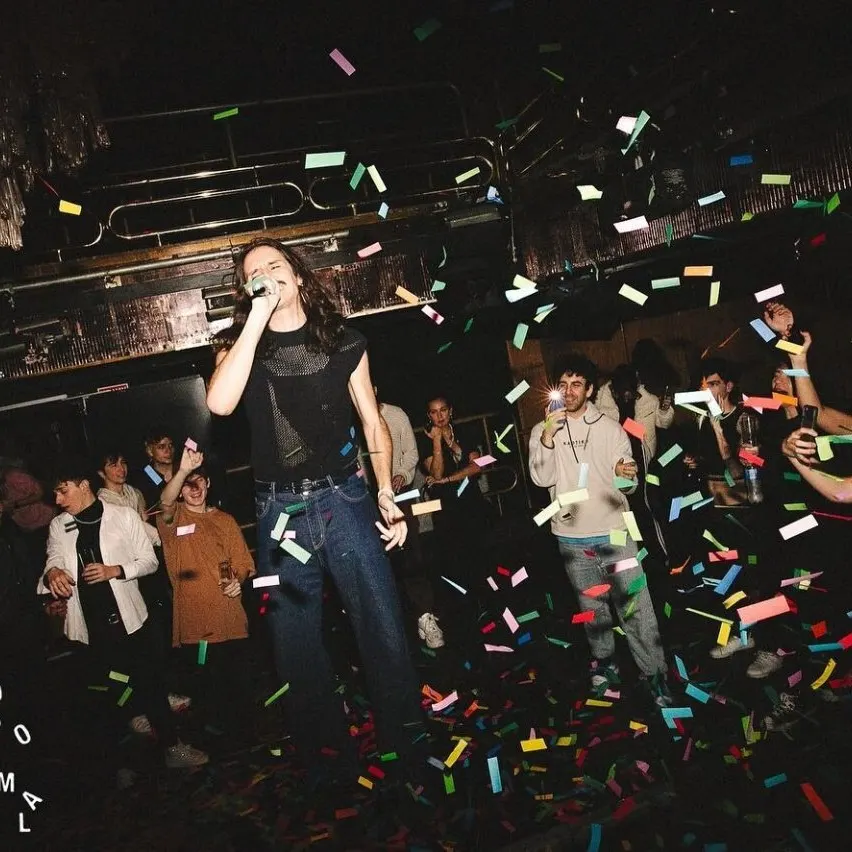 Grupo de personas celebrando en sala oscura con confeti, estructuras metálicas y luces; texto “BORQUE TODO MAL” y “GEN” visible.