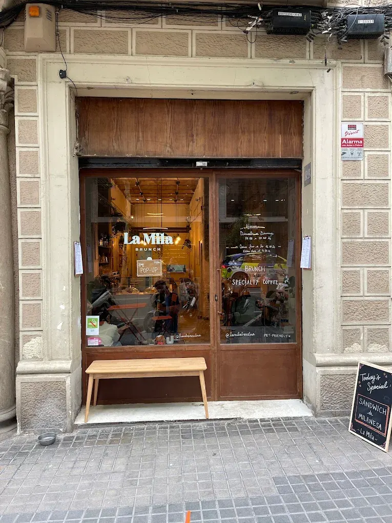 Fachada de La Mila Brunch con puerta de cristal, banco de madera en la entrada y cartel que anuncia el sándwich de milanesa como especial del día.