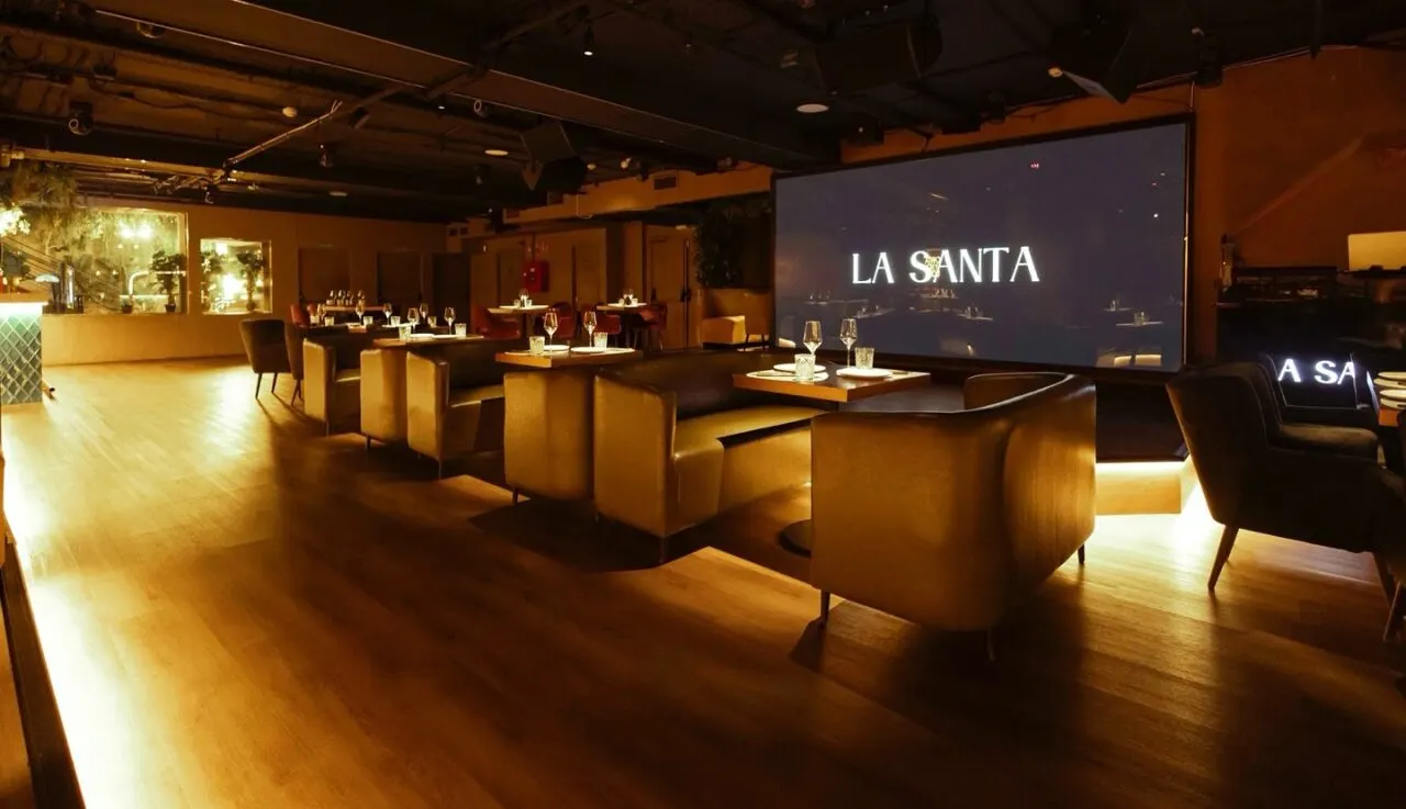 Espacio elegante en La Santa con sofás dorados, mesas con copas de cristal y pantalla iluminada al fondo, todo en una atmósfera cálida y sofisticada.