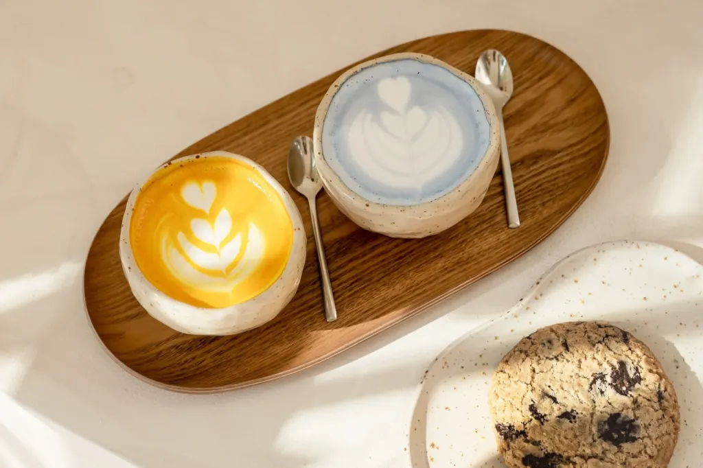 En la imagen se ven dos tazas de cerámica artesanal con latte art, una de color amarillo y otra azul, servidas en una bandeja de madera junto a una cookie con trozos de chocolate.
