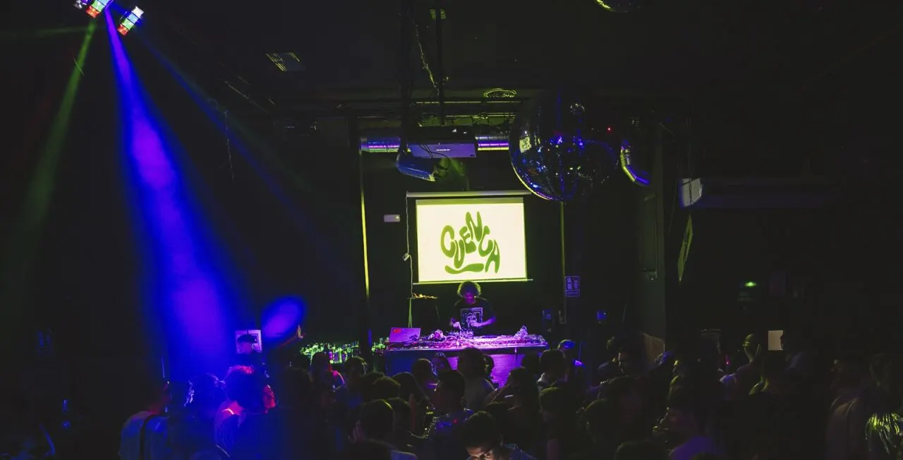 Pista de baile de Cuenca Club llena de gente, con DJ en cabina, luces de colores, bola de espejos y proyector con el logo del club en pantalla.