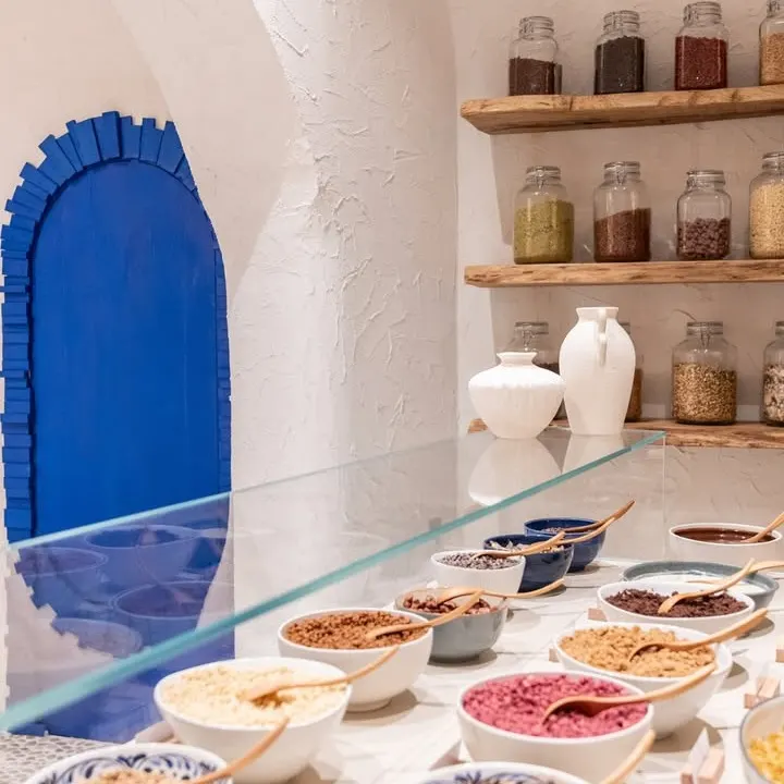 Detalle del mostrador de Myka Ponzano con toppings en cuencos, estanterías rústicas y una puerta azul que contrasta con las paredes blancas.