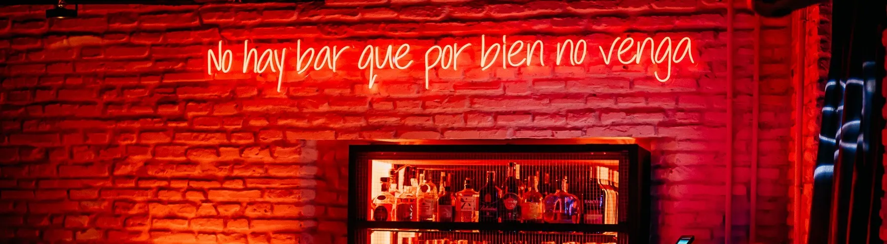 Bar Dadá iluminado en tonos rojos con un neón que dice "No hay bar que por bien no venga" y una barra con botellas de licor al fondo.