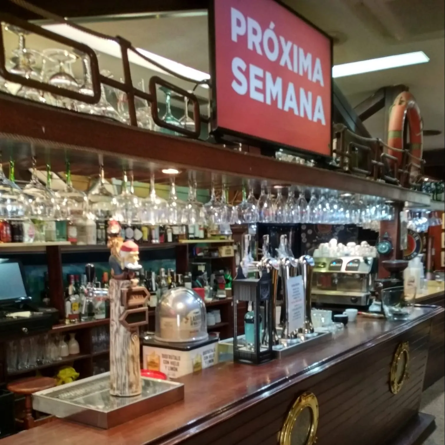Barra de madera con detalles náuticos, cristalería colgante, grifos de cerveza y un cartel luminoso que anuncia "Próxima semana".