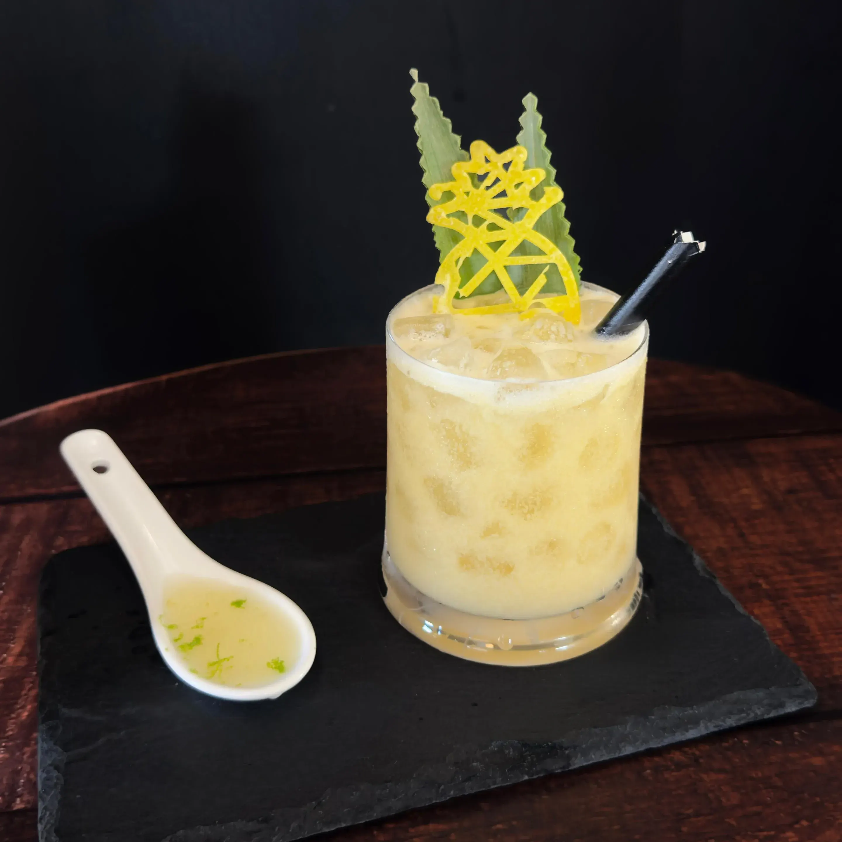 Cóctel cremoso con hielo en vaso bajo, decorado con piña y hoja, sobre pizarra negra; cuchara con líquido al lado en mesa de madera.