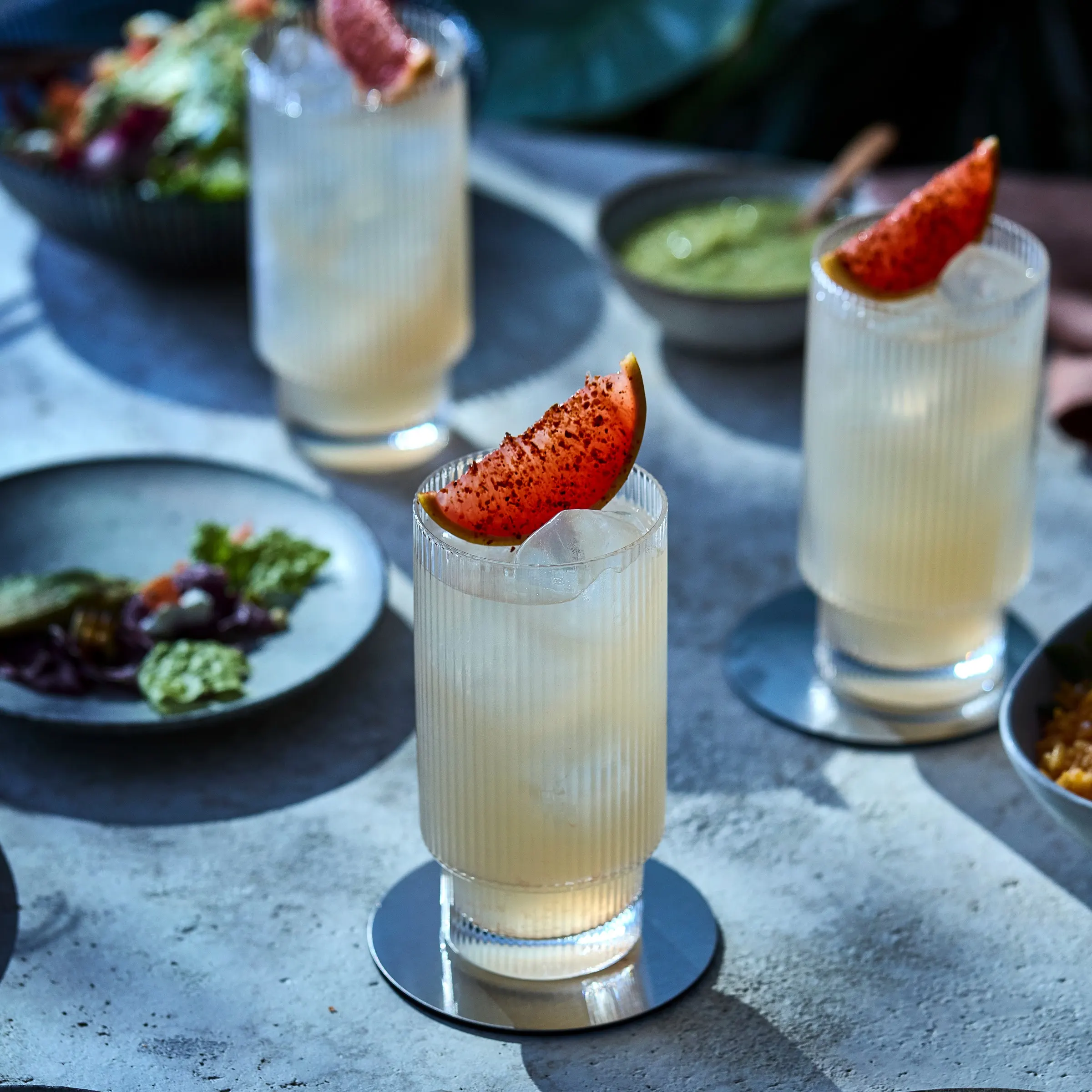 Tres vasos de paloma con hielo y una rodaja de pomelo rociada con chile en polvo, en una mesa de comedor con platos de ensalada y salsas, preparados para una comida