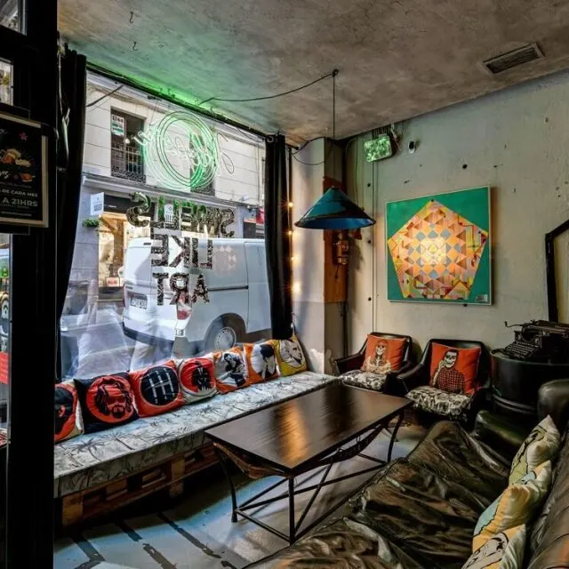 Interior de café pequeño con sofás, cojines coloridos, arte abstracto, cartel de neón en ruso y furgoneta blanca visible por la ventana.