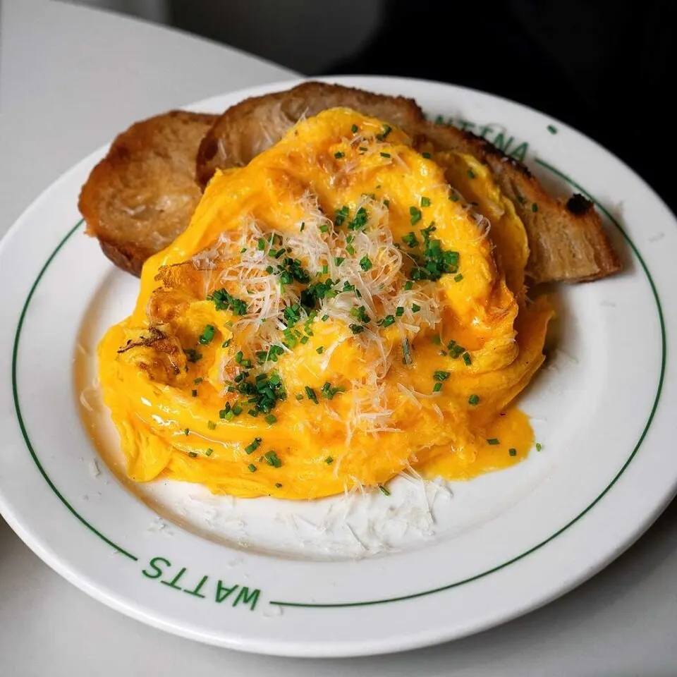 Huevos revueltos con cebollino y queso rallado, acompañados de dos tostadas. El plato tiene inscrito el nombre “WATTS”.