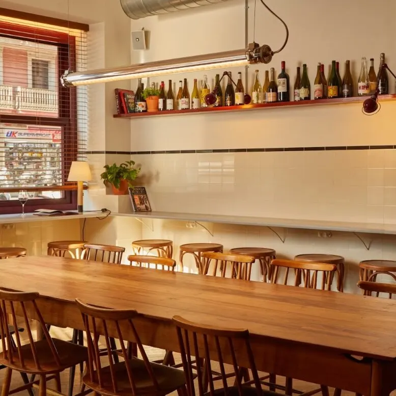 Café acogedor con mesa larga de madera, estantería con botellas, lámparas colgantes, taburetes y ventana con vista urbana.