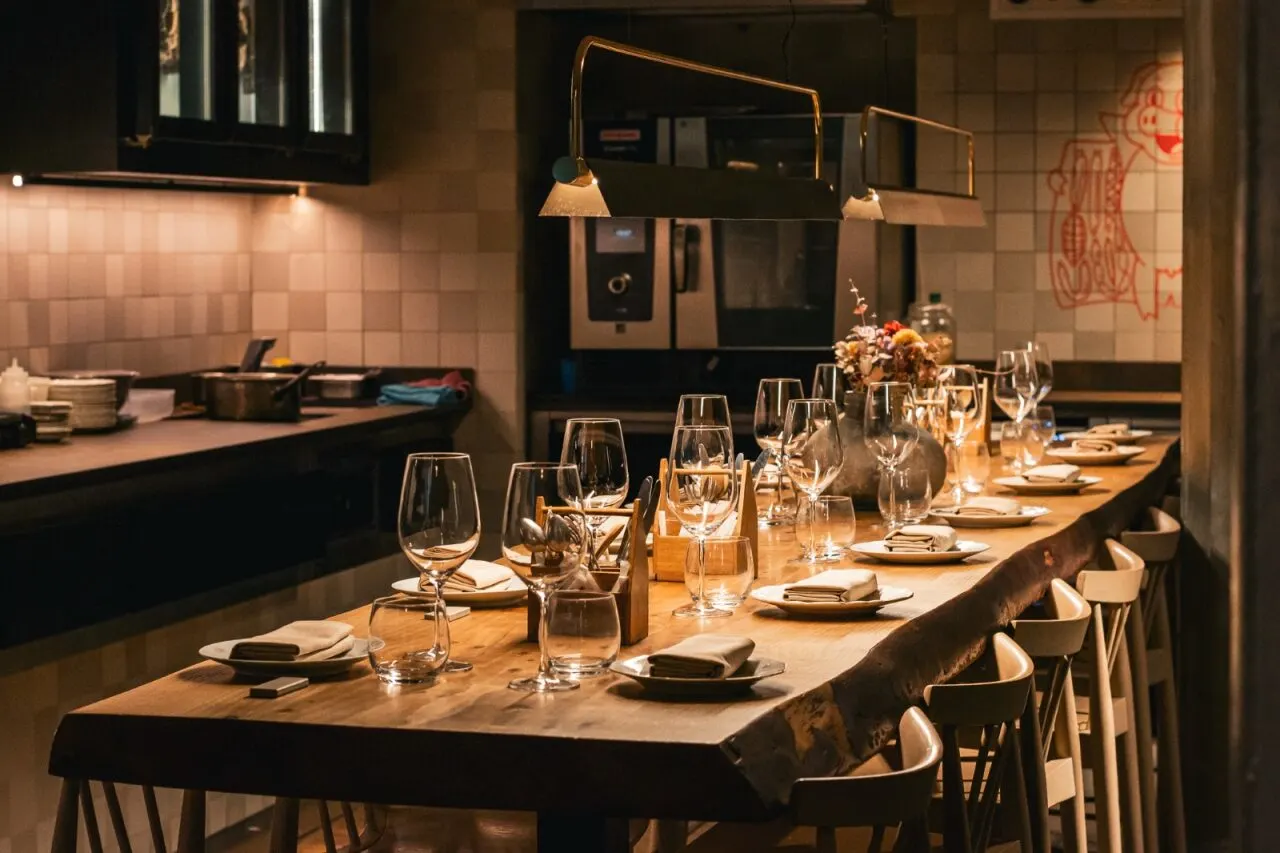 Mesa de comedor bien puesta con copas, cubiertos y flores. Fondo con cocina equipada, paredes de azulejos y luces colgantes. Ambiente cálido.