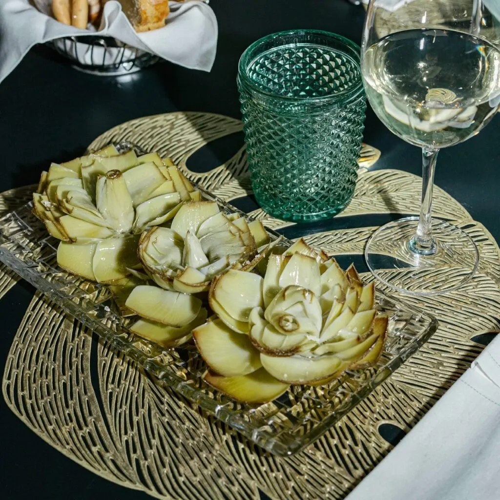 Mesa con plato de alcachofas, vaso verde texturizado, copa de vino blanco y cesta con pan y colines sobre mantel decorativo.