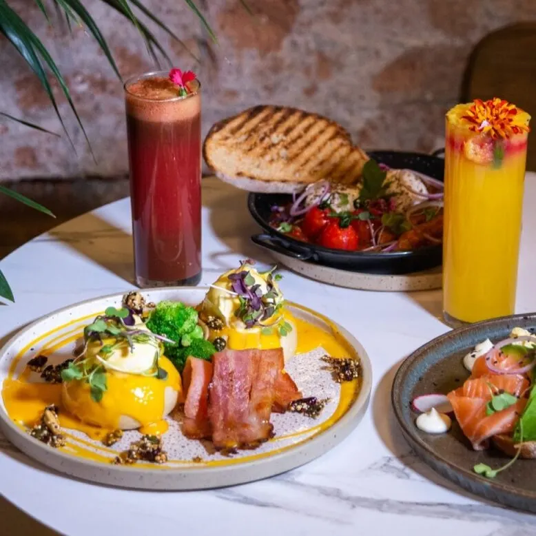 Mesa con huevos benedict, salmón con crema, ensalada con pan plano, zumos rojo y naranja decorados, fondo de ladrillo y hojas verdes.
