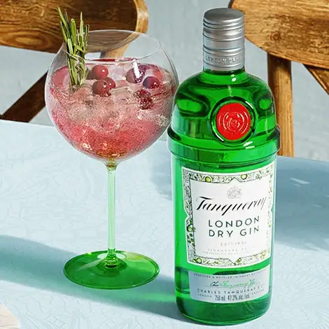 Copa de gin tonic rosa con hielo y bayas, acompañada de una botella de Tanqueray London Dry Gin en una mesa con decoración floral