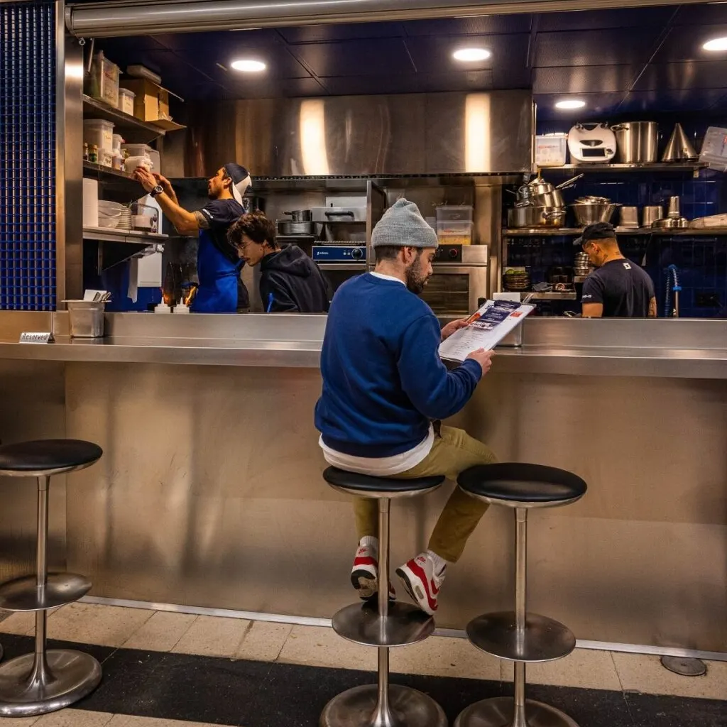 Persona sentada en taburete leyendo menú en barra de restaurante. Tres empleados trabajan en cocina con estanterías y electrodomésticos de acero.