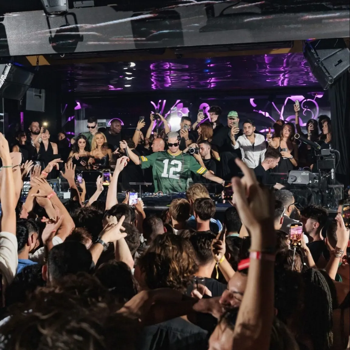 Club lleno con público levantando manos y móviles; DJ con camiseta verde “12” en cabina, luces púrpuras y personas detrás del escenario.