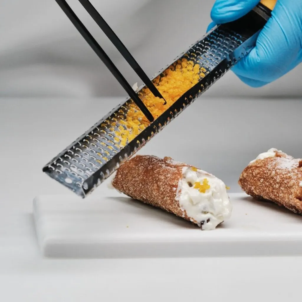 Manos con guantes azules rallan cítrico sobre dos cannoli con crema en plato blanco rectangular; decorados con ralladura por encima.